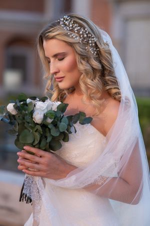 A bride in a white dress holds a eucalyptus and white rose bouquet while wearing a crystal headpiece and veil.