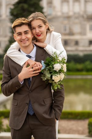 Romantic wedding portrait of couple embracing outdoors with bride holding white bouquet near palace grounds and pond.