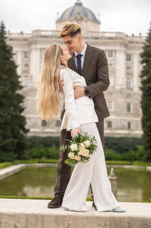 A romantic couple in formal attire embraces near a grand historic building with a domed roof and reflecting pool.