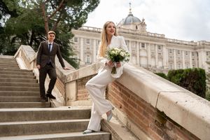 Wedding couple playfully poses on grand stone steps in front of ornate historical palace building with bouquet.