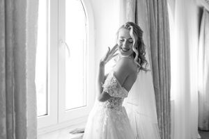 Black and white photo of a bride in a strapless lace wedding gown standing by a bright window.