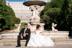 Elegant wedding couple sits at ornate fountain in front of grand palace with classical architecture and manicured gardens.