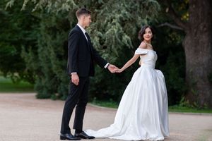 A couple in formal wedding attire dances together in a park setting with evergreen trees in the background.