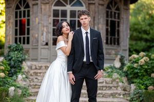 Couple in formal wedding attire poses together on stone steps of a rustic building in natural outdoor setting.