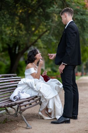 A couple in wedding attire shares a playful moment on a park bench during their outdoor photo session.
