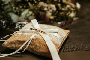 A rustic burlap wedding ring pillow decorated with white ribbons and delicate beading in soft lighting.
