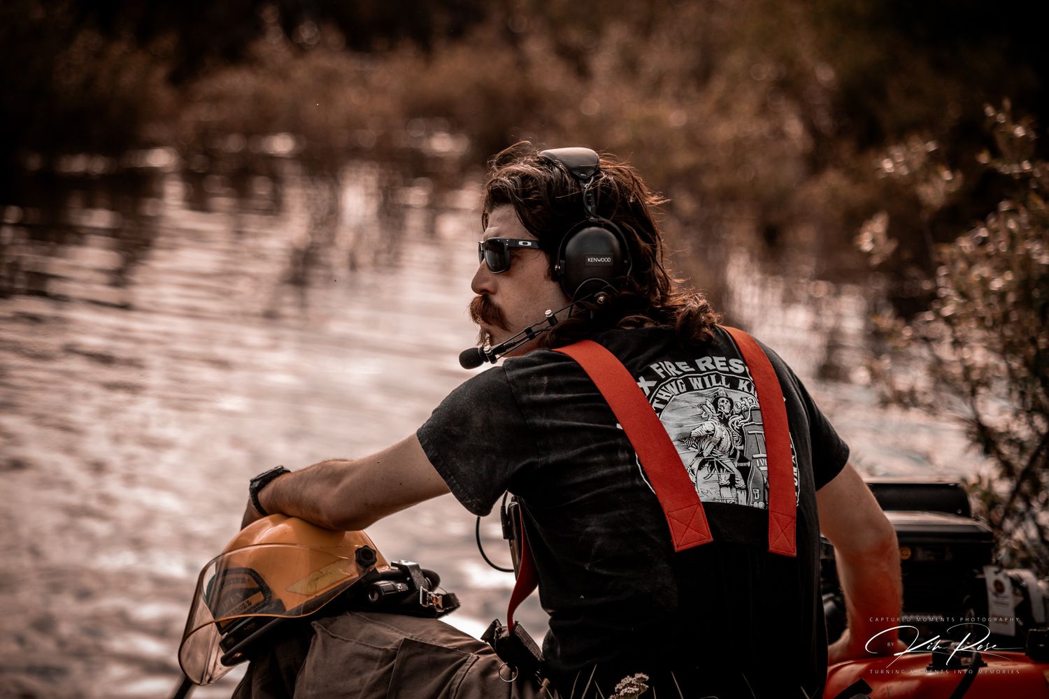 Man with headphones on a boat near a river, wearing a black graphic tee and red suspenders.