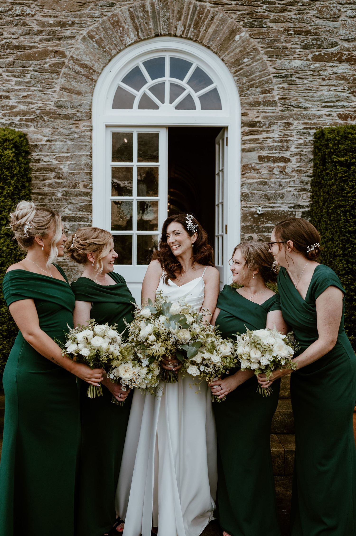A wedding party in emerald green dresses and white gowns gather against a rustic stone wall with arched windows.