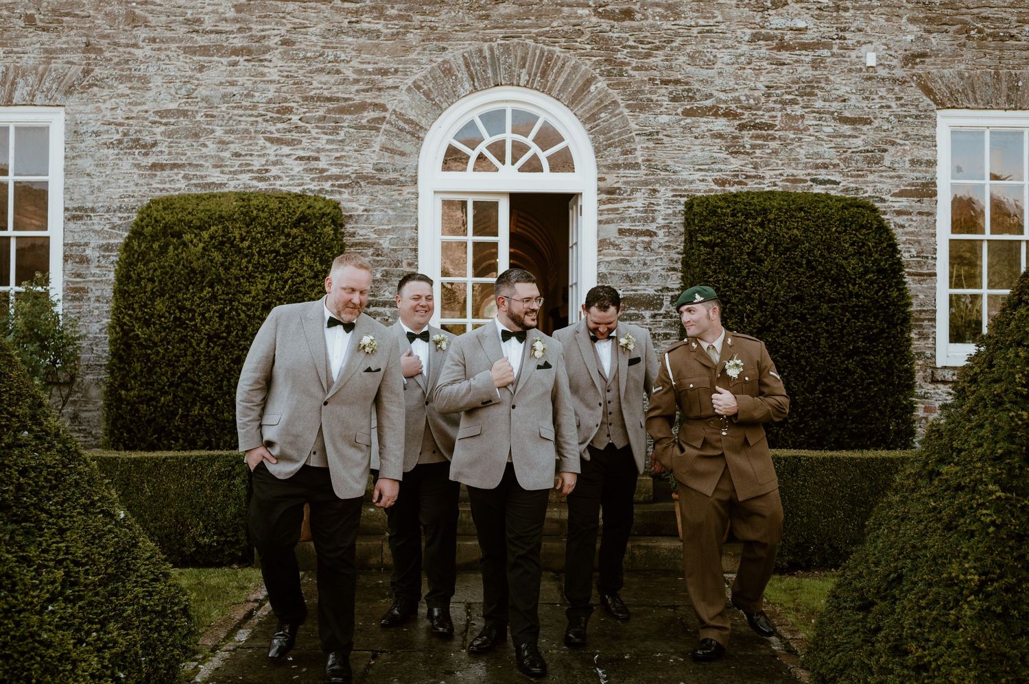 A wedding party in gray suits stands together laughing outside a stone building with arched doorway and manicured hedges.