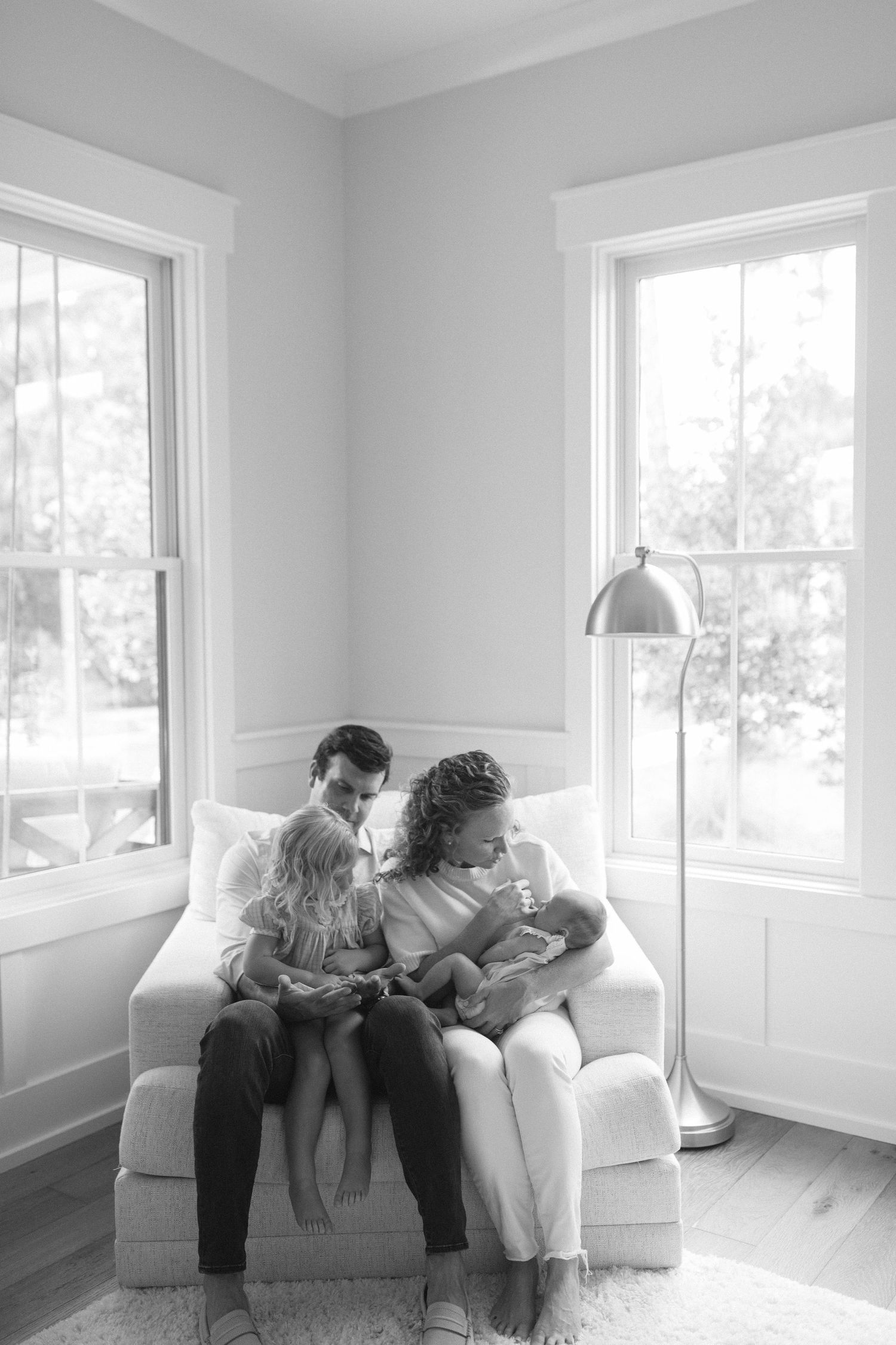 Black and white photo by B. Logsdon Photography of a couple sitting together on a white armchair in a bright corner room with two windows.