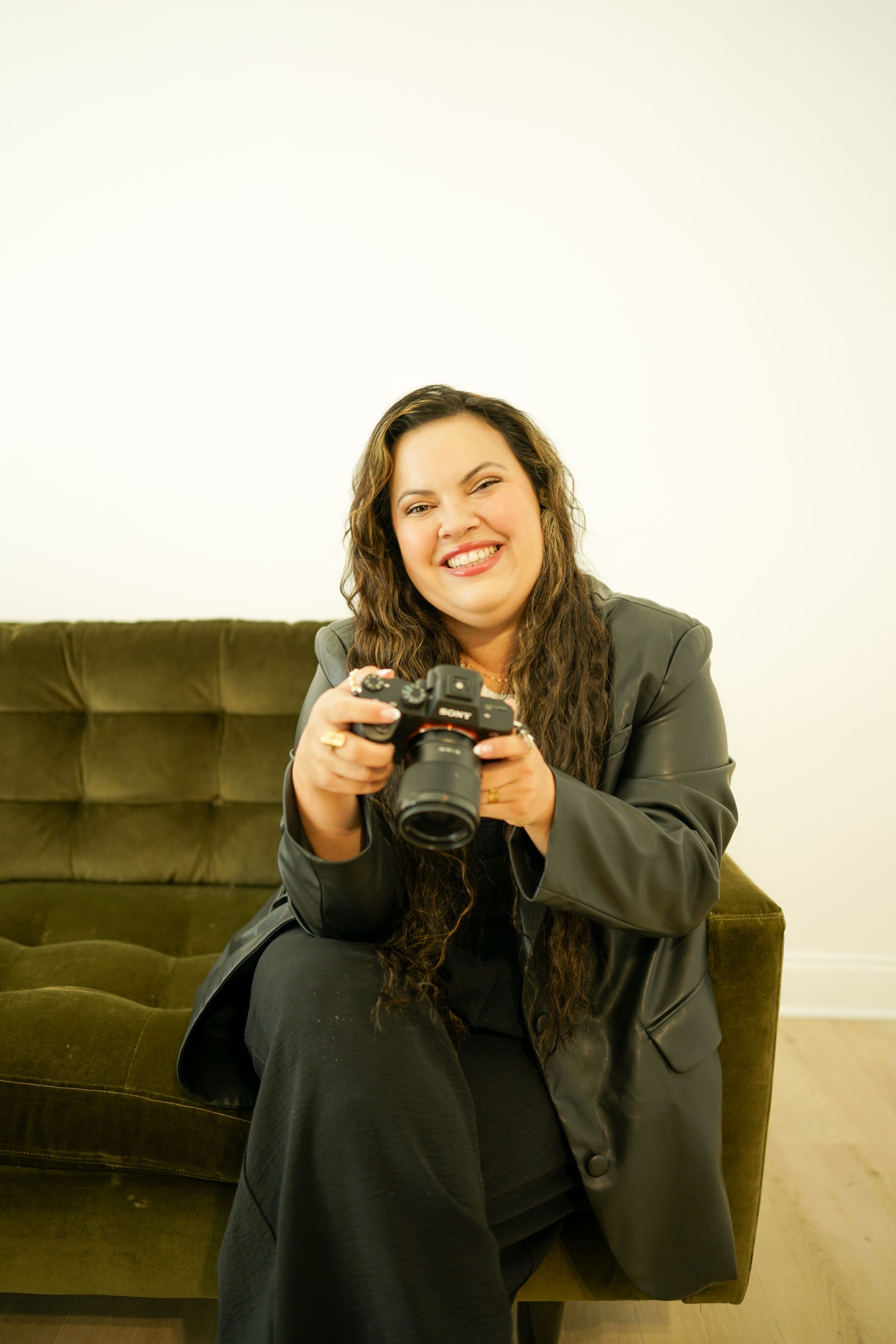 Photographer with camera smiling and sitting on brown couch in indoor setting.