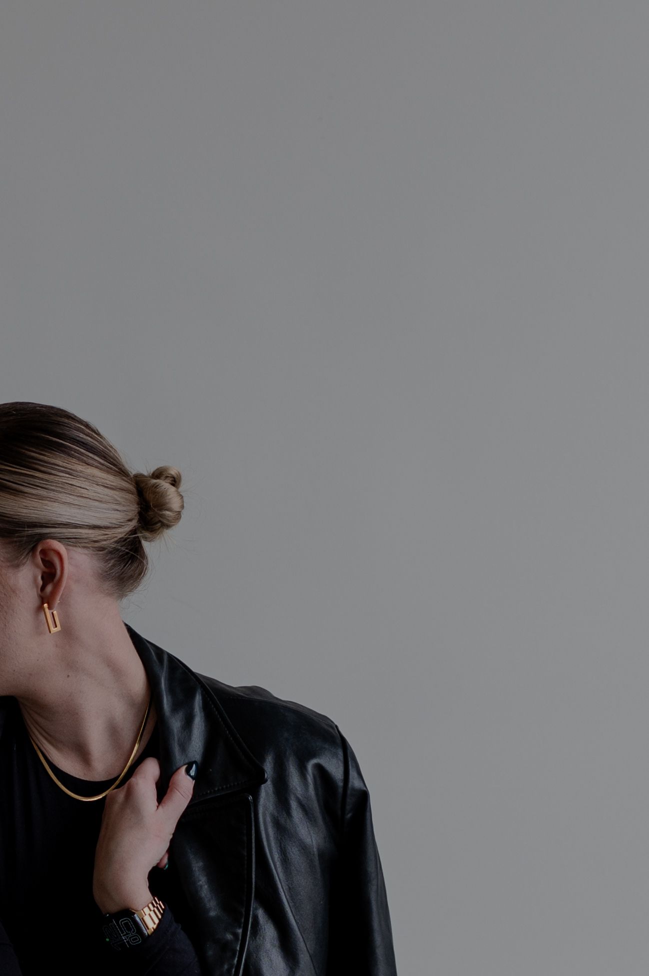 Side profile of person wearing black leather jacket and gold jewelry against grey background.
