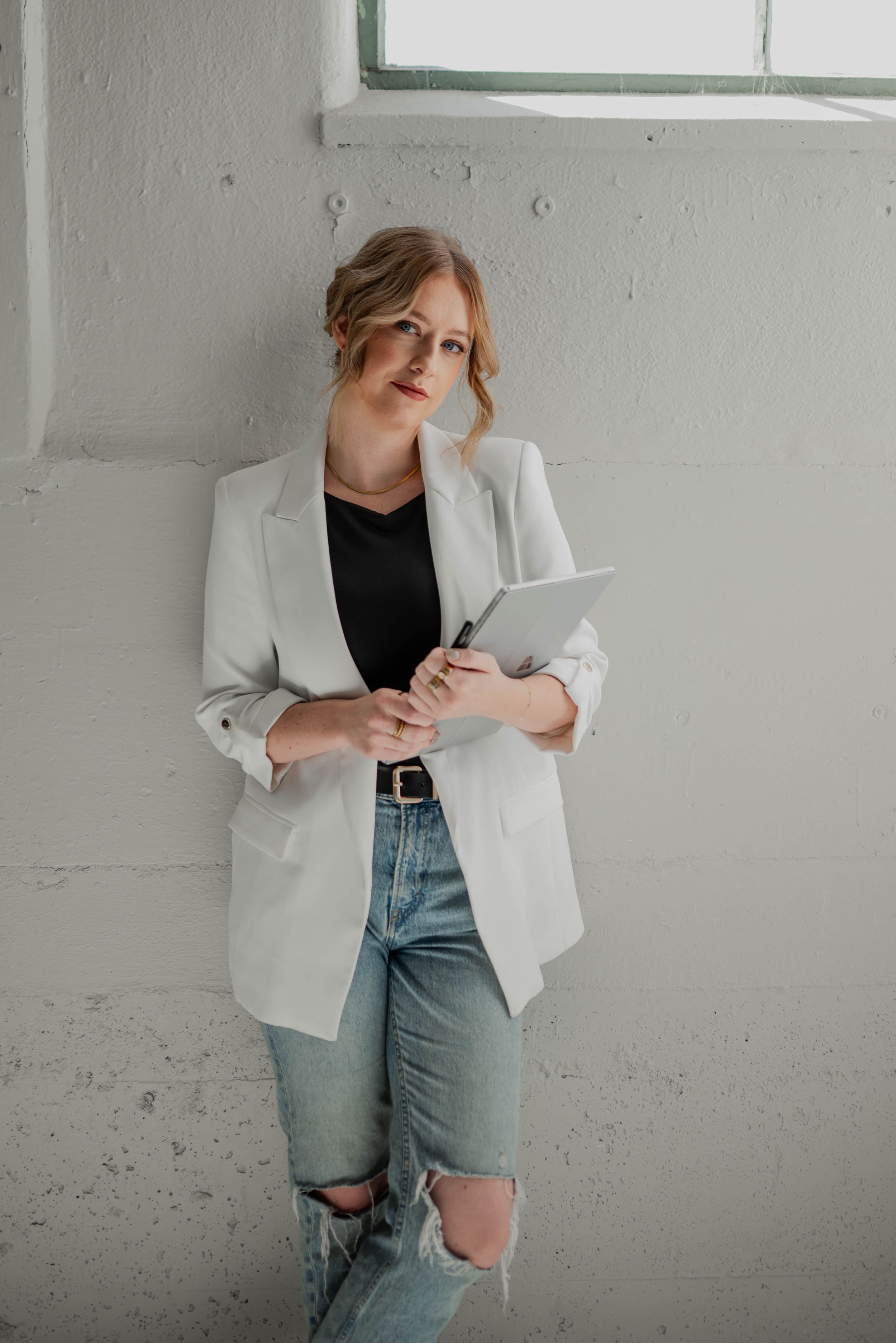 Fashion portrait of model in white blazer and distressed jeans against industrial wall.