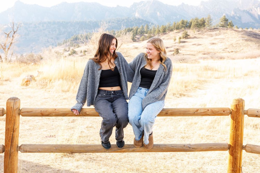 Two people sit together on a wooden fence with scenic mountain views in the background on a sunny day.