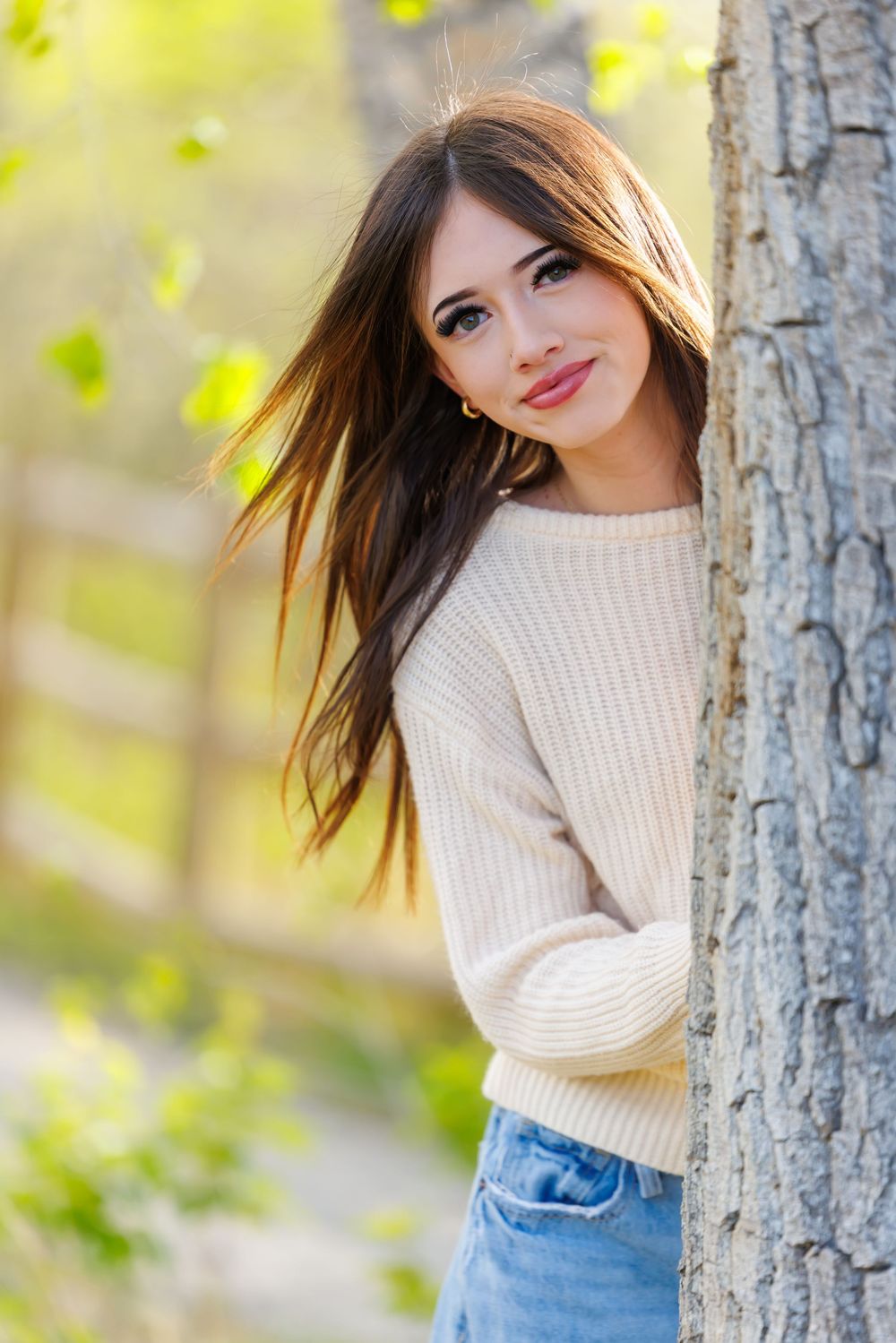 Young brunette woman in cream sweater and jeans leaning against tree trunk in sunny green park setting.