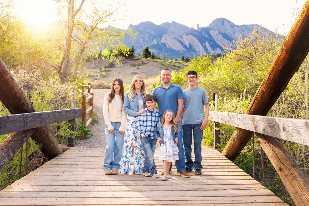 Family of six posing on a wooden bridge with Flatirons mountains and golden sunlight in the background.