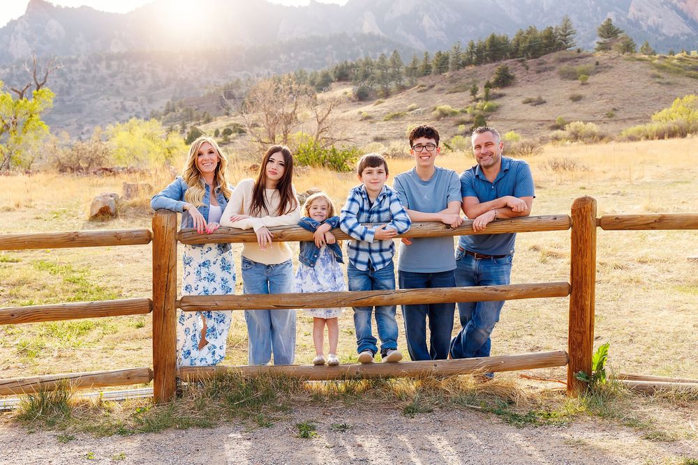 Family of six posing at a wooden fence with Rocky Mountain foothills backdrop on a sunny day.