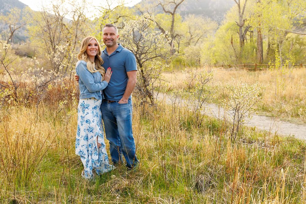 A smiling couple poses together in a scenic outdoor field with golden grasses and green trees in the background.