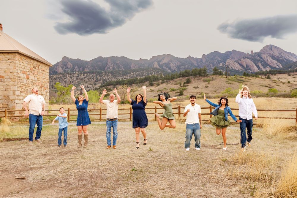 Family jumping joyfully outdoors with Rocky Mountain backdrop, rustic fence, and dramatic cloudy skies in Colorado.