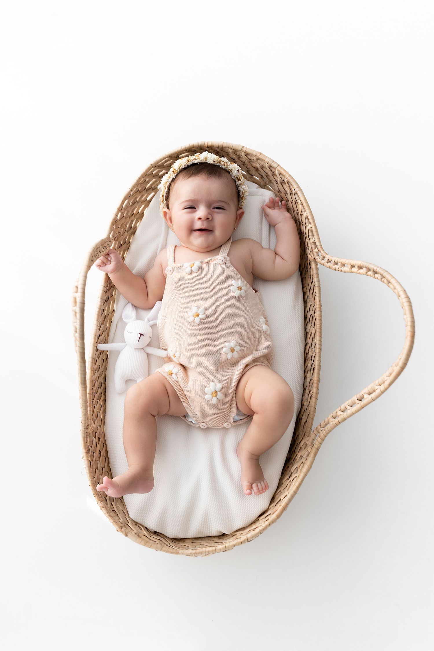 Baby wearing floral romper lies contentedly in woven bassinet during portrait session.