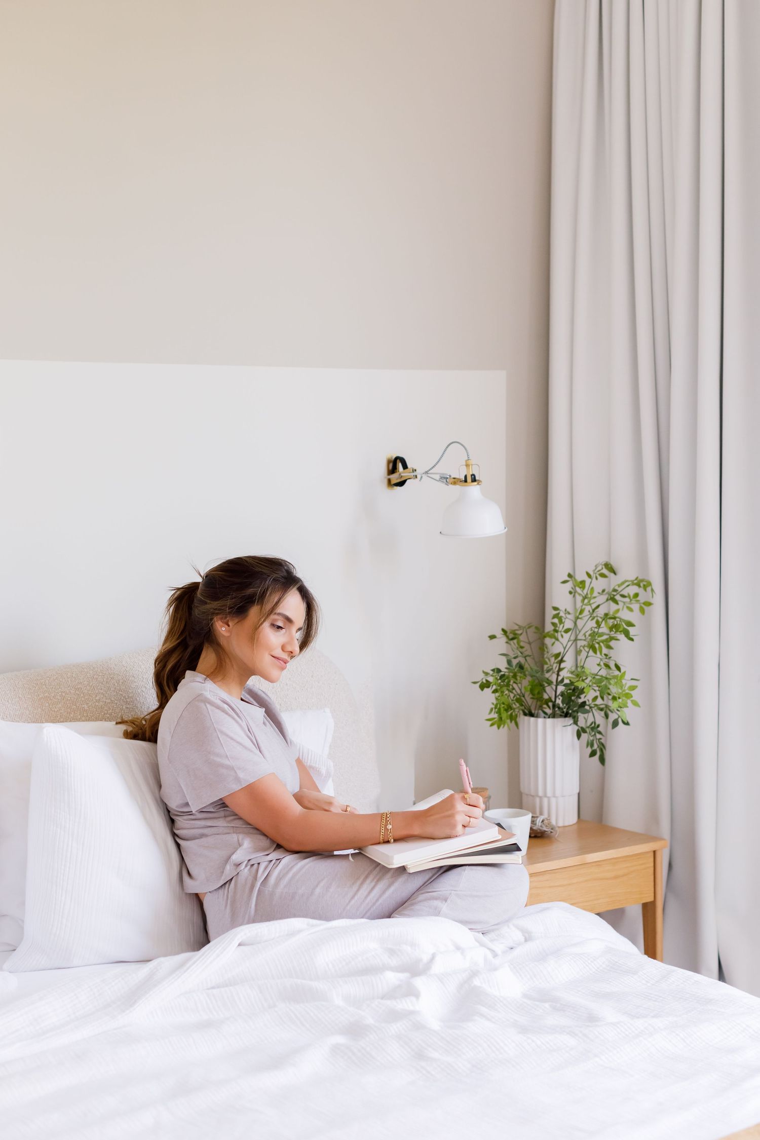 Someone enjoying a peaceful morning while journaling in bed next to a potted plant and white curtains.