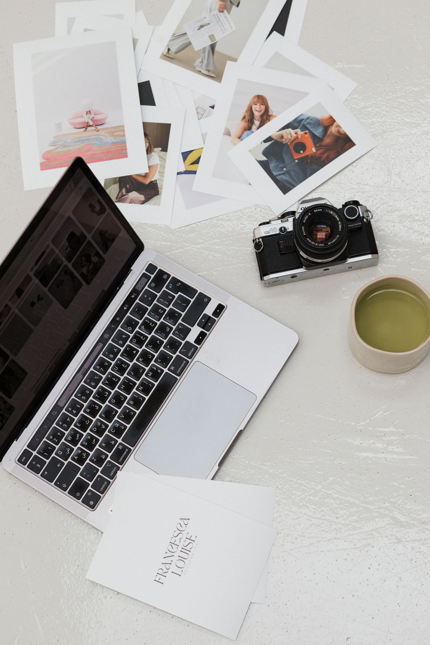 Branding photographer set up. Laptop keyboard with scattered photographs and a matcha latte on white marble desk surface.