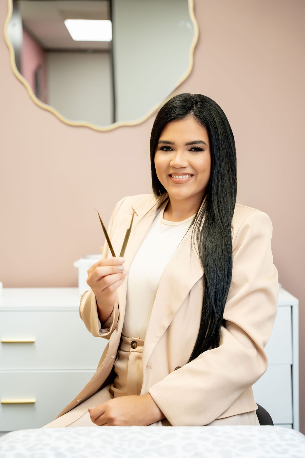 Professional beauty technician in beige blazer smiling while holding beauty tools in a salon setting.