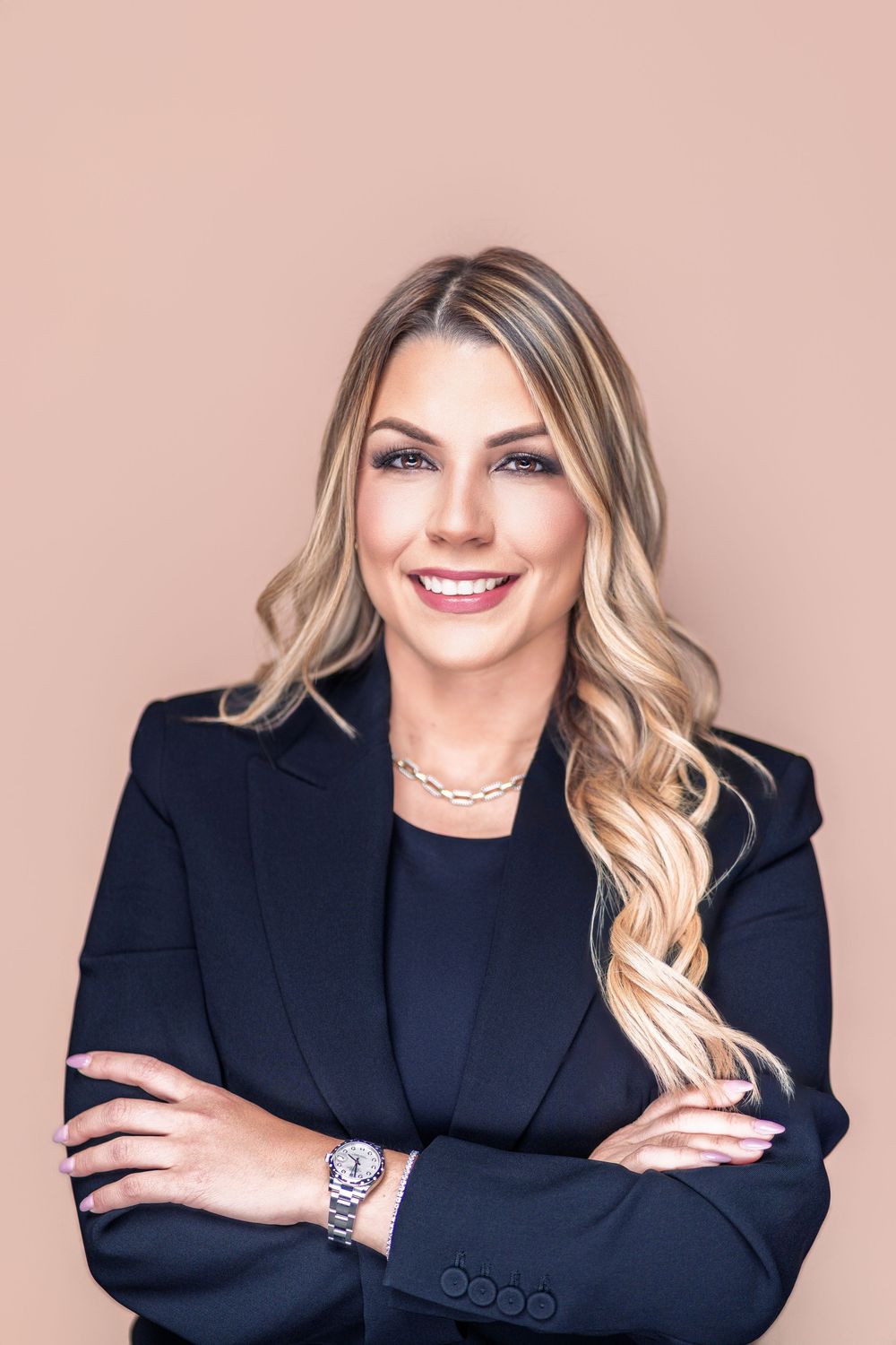 Professional headshot of a person in a navy blazer with blonde wavy hair and jewelry against a pink background.