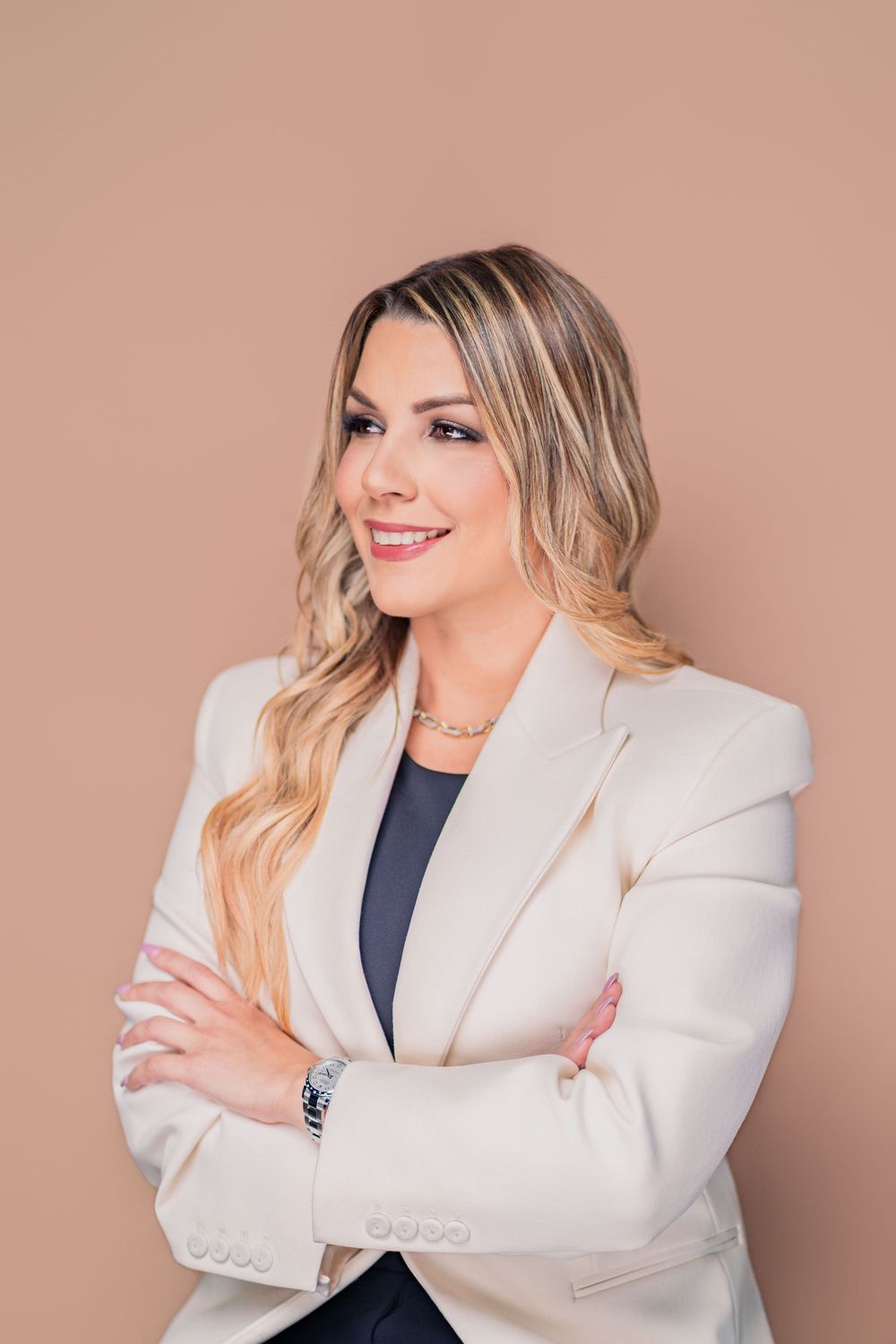 Professional headshot of a person in a cream blazer and dark top smiling with arms crossed against a neutral background.