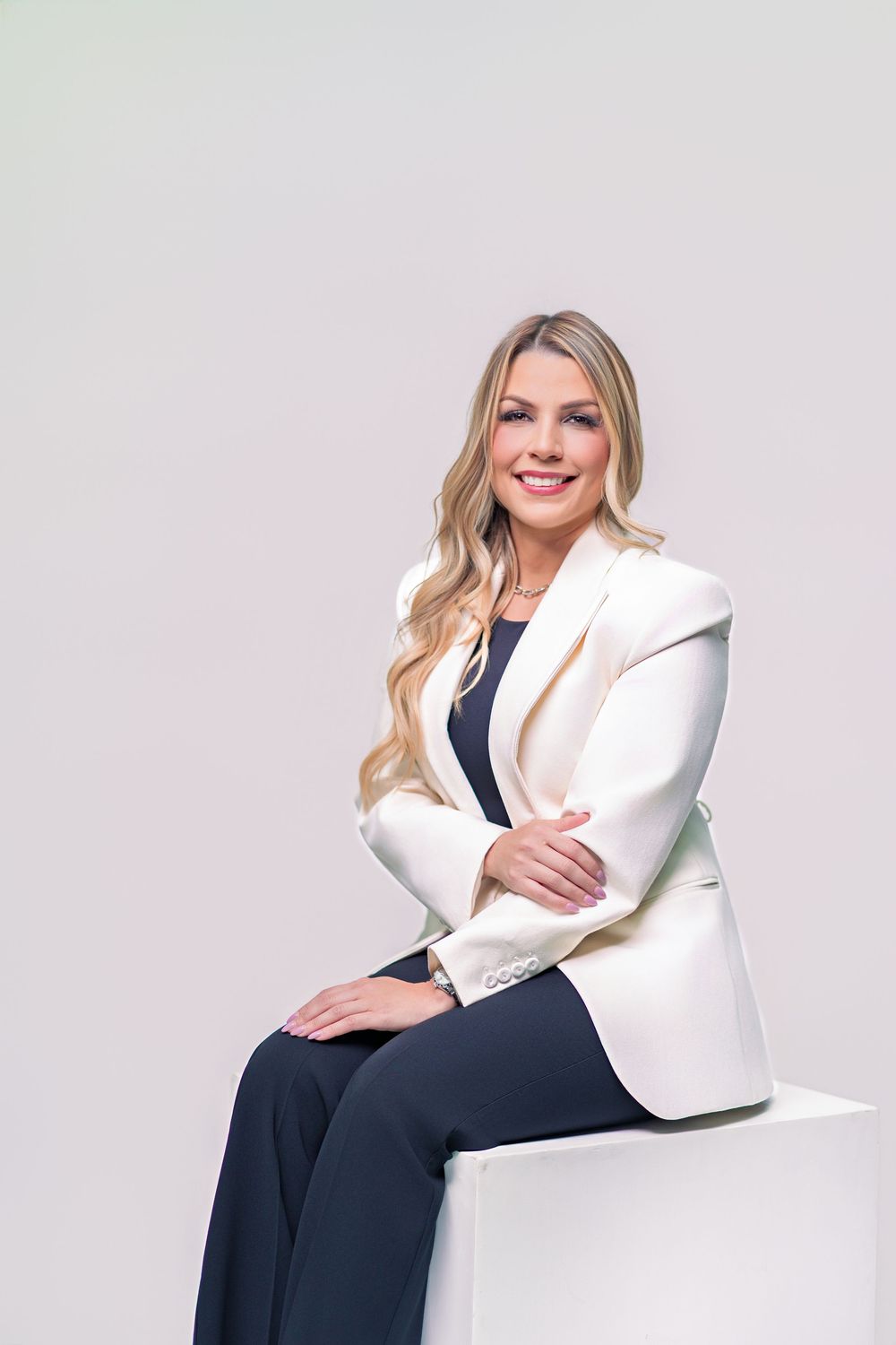 Professional business portrait of someone in a white blazer and navy dress sitting elegantly against a light background.