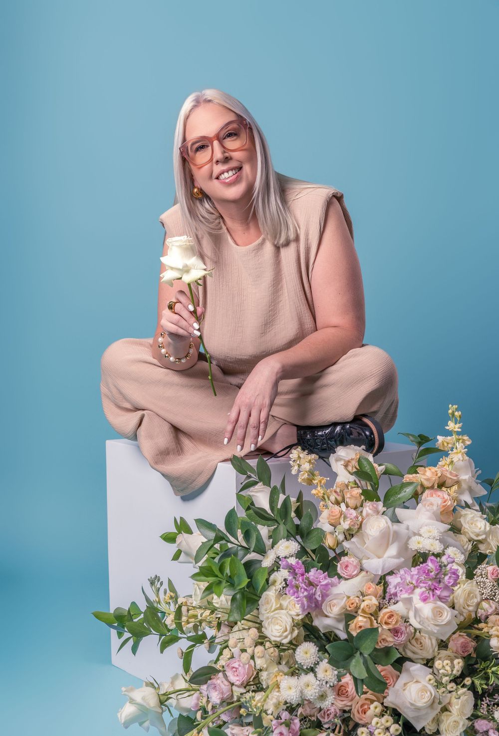 Elegant professional seated on white cube surrounded by floral arrangements with beverage.