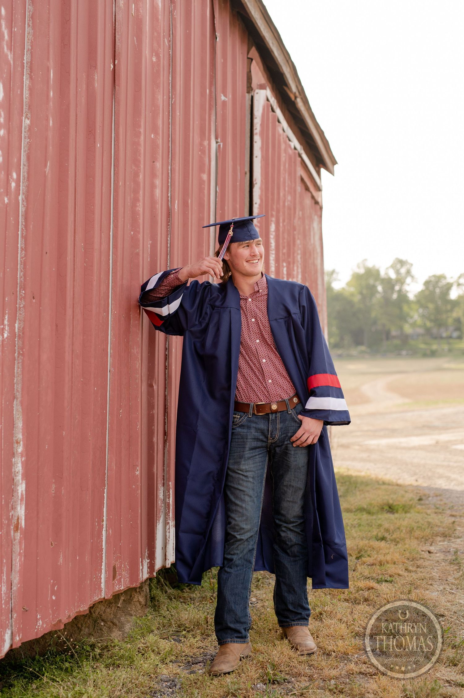 Graduate in navy cap and gown poses confidently against red barn wall.