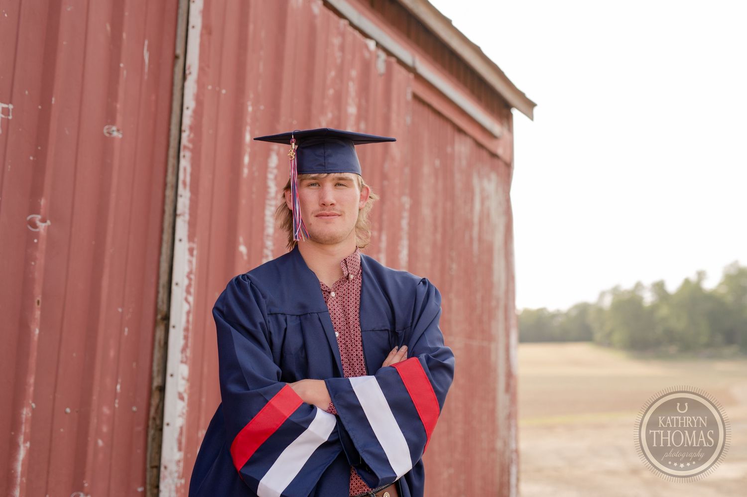 Graduate in navy blue graduation gown poses against weathered red barn wall.
