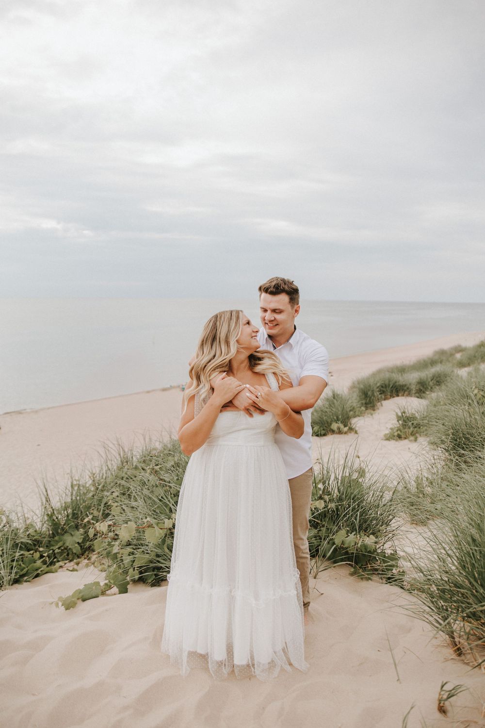 A couple in white outfits shares romantic moments among beach grass.