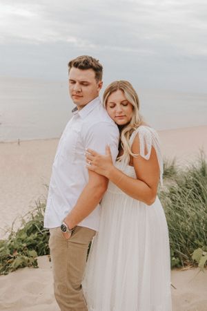 A couple in casual attire embraces on sandy dunes near the ocean.