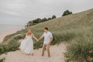 A couple walks hand in hand along grassy coastal dunes.