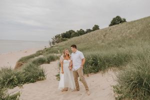 A couple walks together on a sandy beach path with grassy dunes and water in the background.