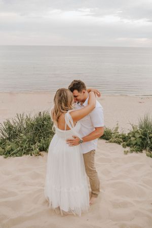 A romantic embrace on a sandy beach with gentle waves and coastal vegetation in the background.
