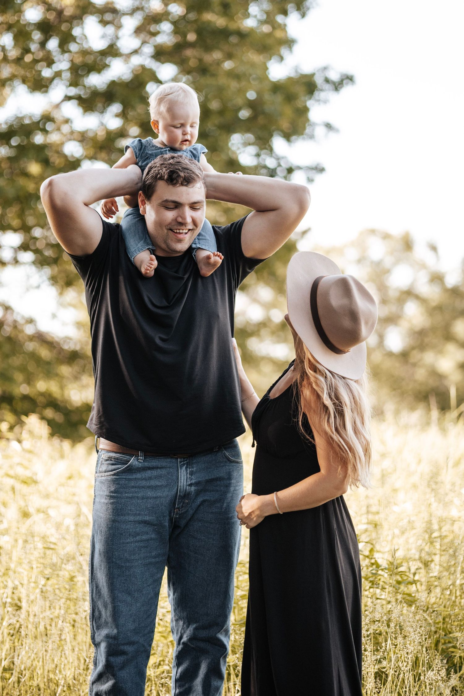 A baby rides on shoulders while a couple enjoys a sunny afternoon in a grassy field.
