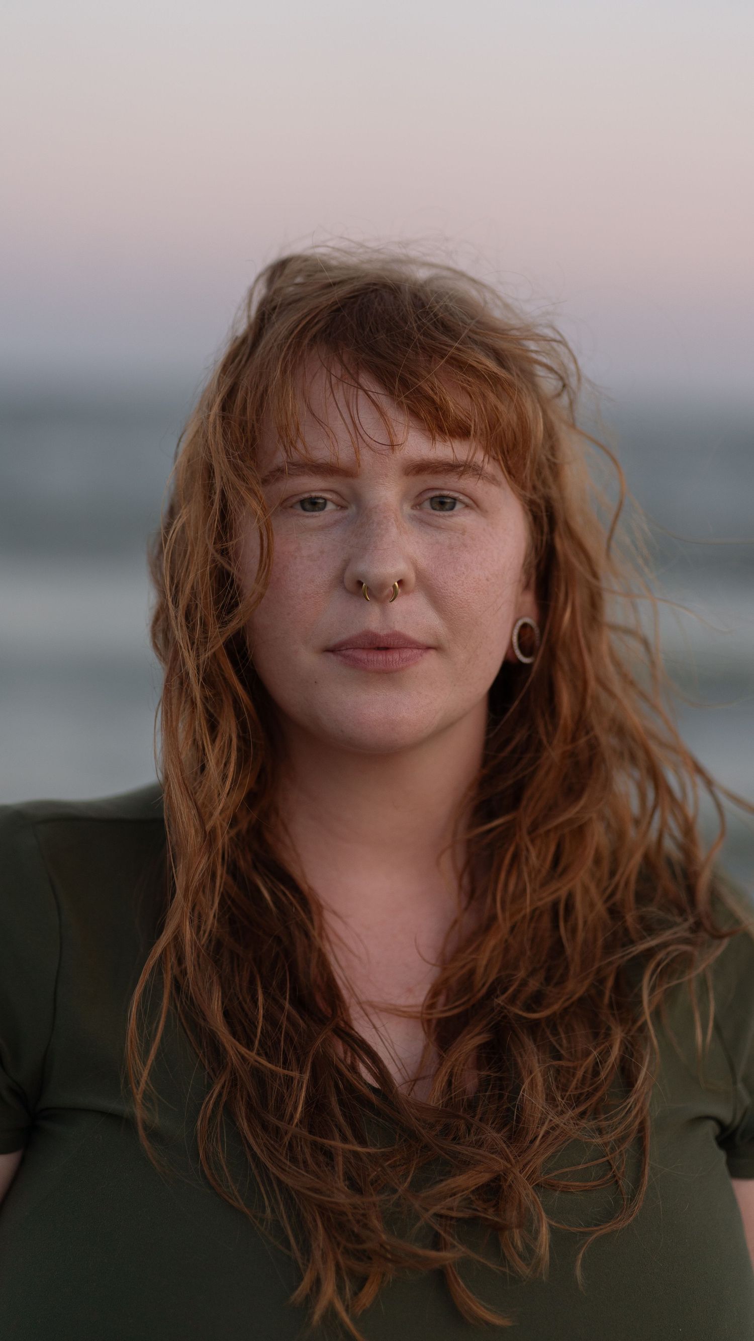 Portrait in soft evening light, a woman with long wavy red hair and green top against blurred coastal background.