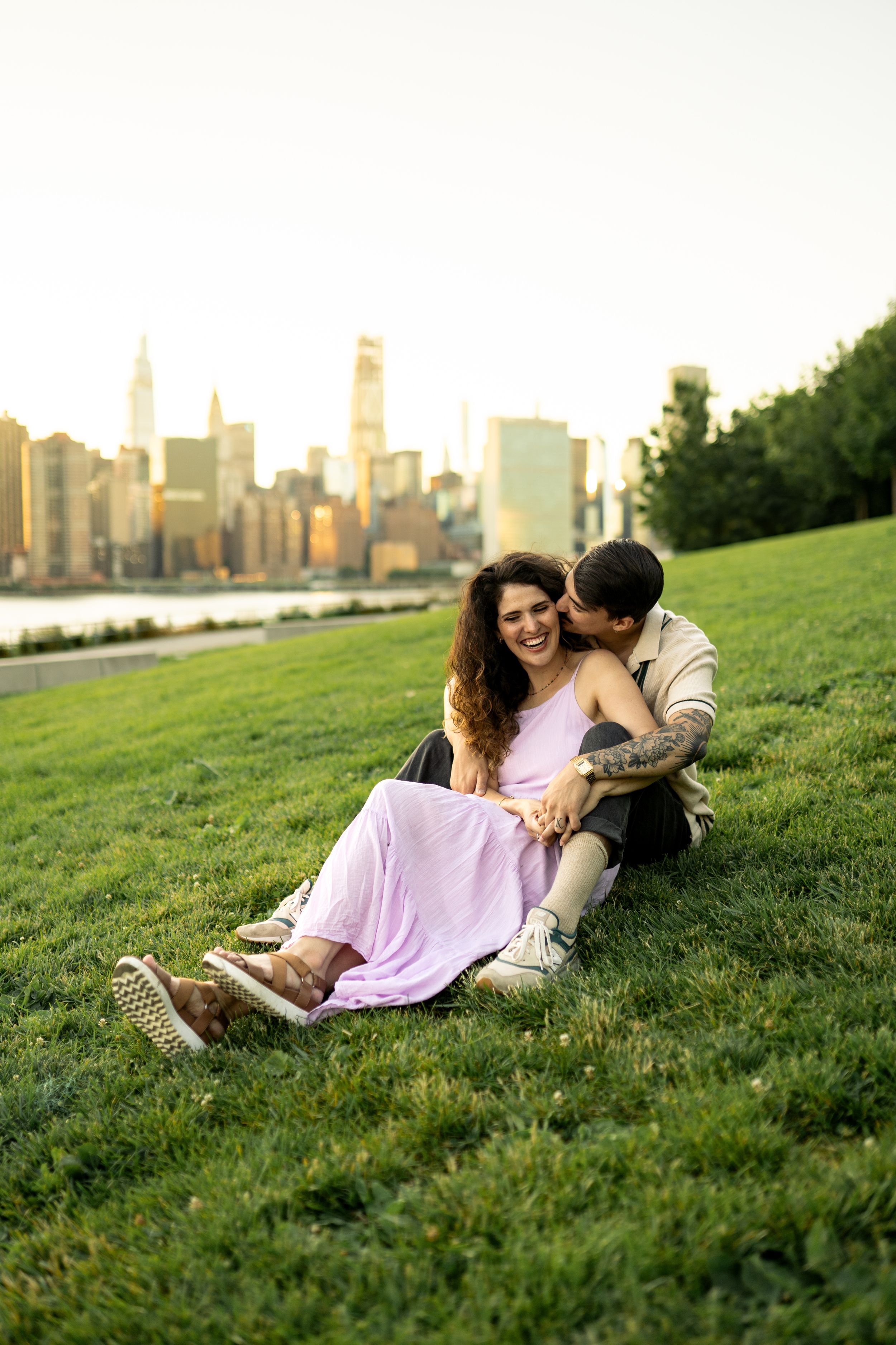 A couple shares a playful moment on grass with the Manhattan skyline visible in the distance.