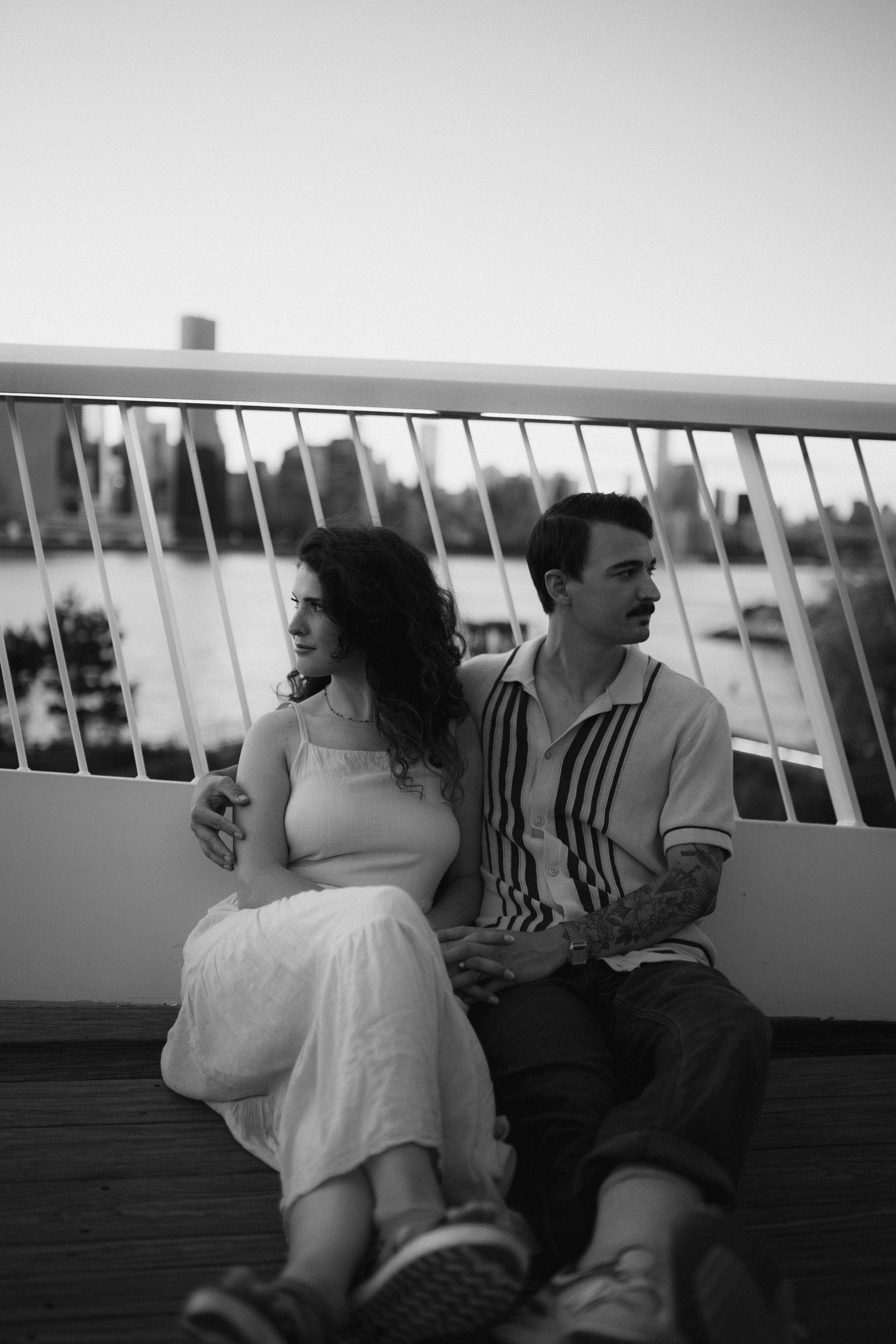 Black and white photo of couple sitting casually on waterfront deck.
