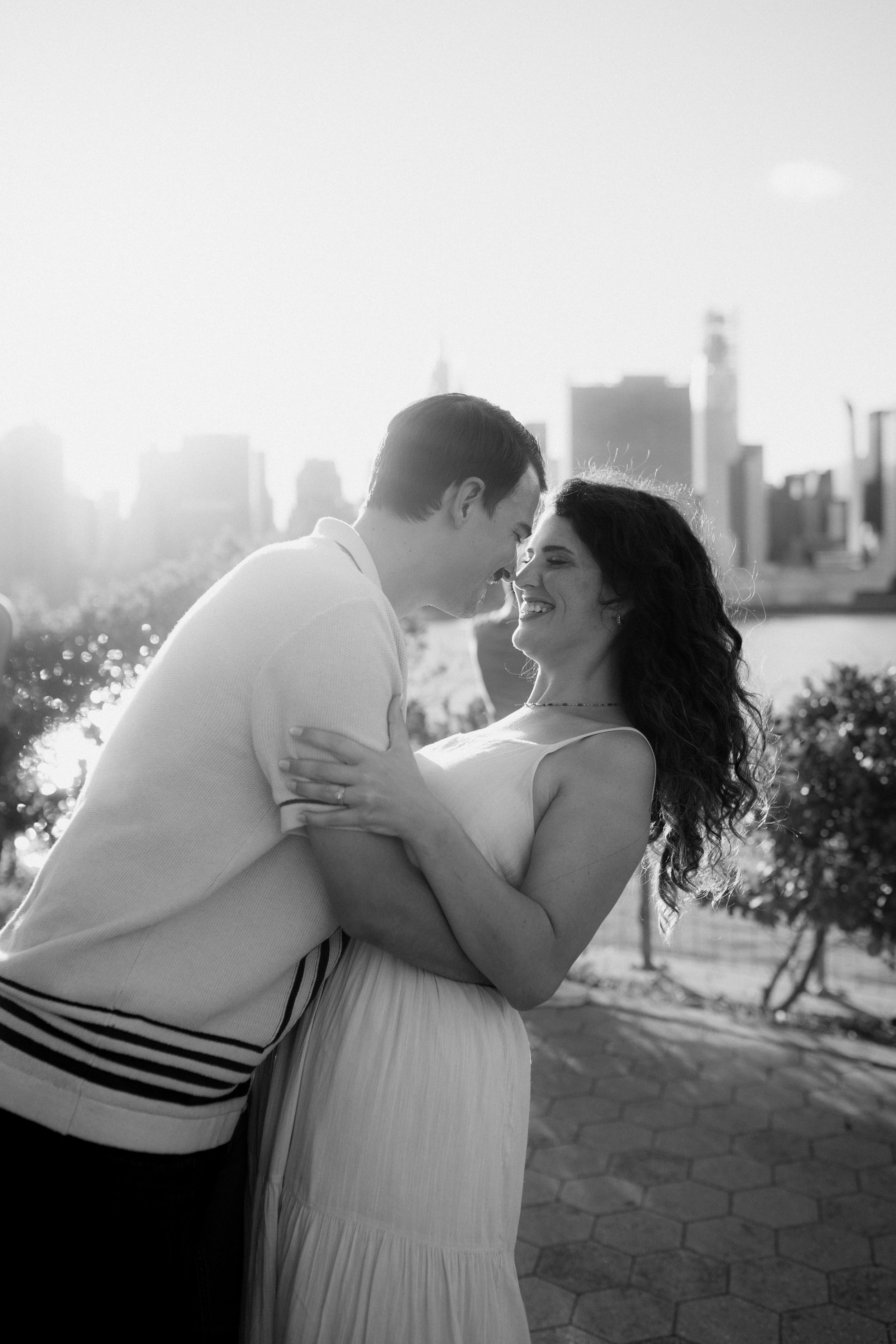 Black and white photo of a couple sharing a romantic moment with city skyline behind them.