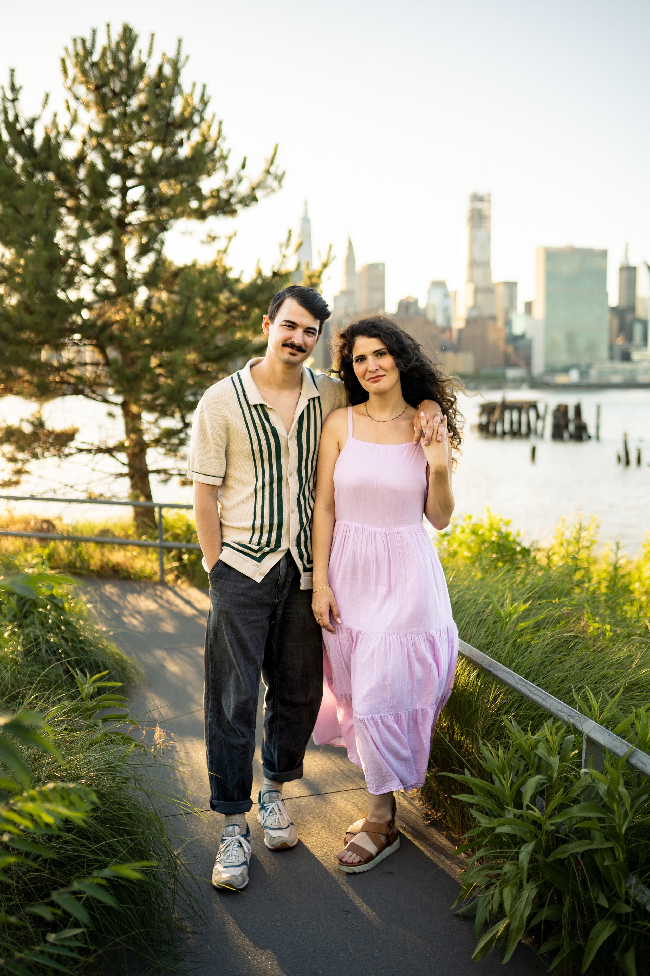 A couple walks along a waterfront path with the city skyline visible in the background.