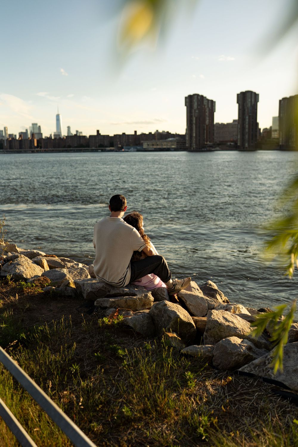 A couple sits on rocks by the waterfront at sunset overlooking the New York City skyline.