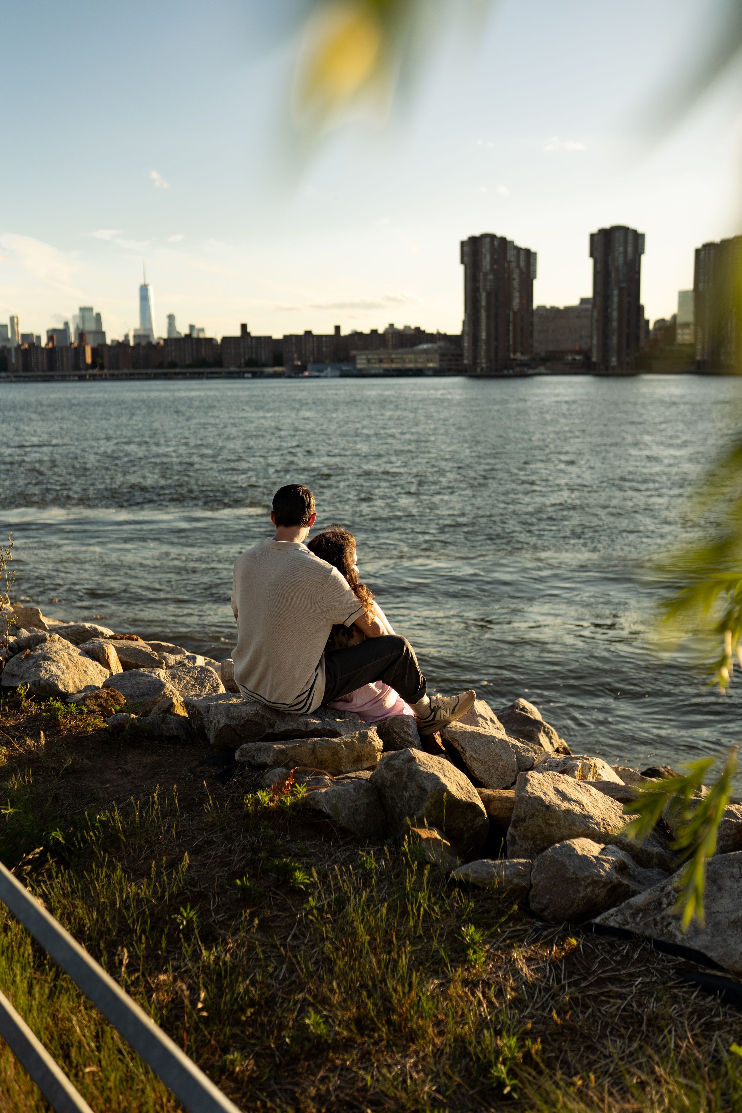 A couple sits on rocks by the waterfront at sunset overlooking the New York City skyline.