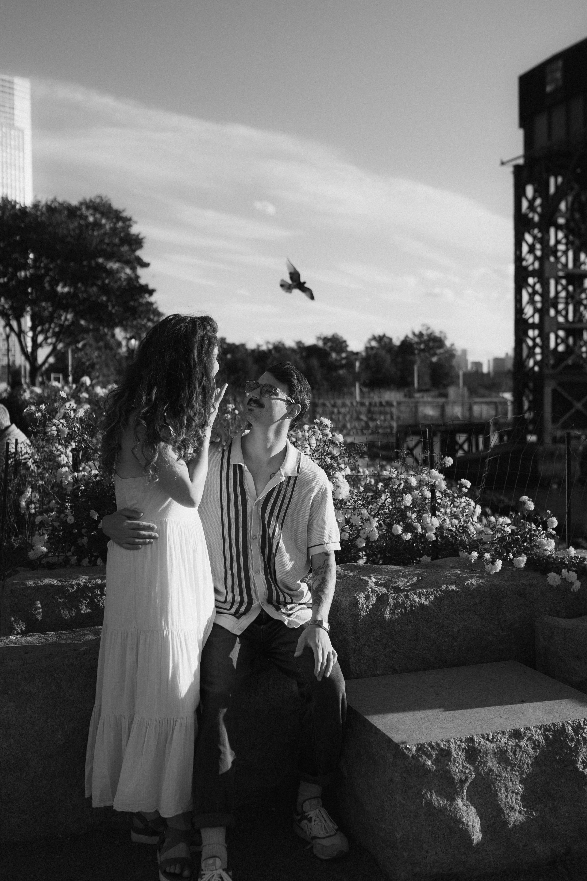 Couple in vintage attire poses in garden setting with city backdrop.