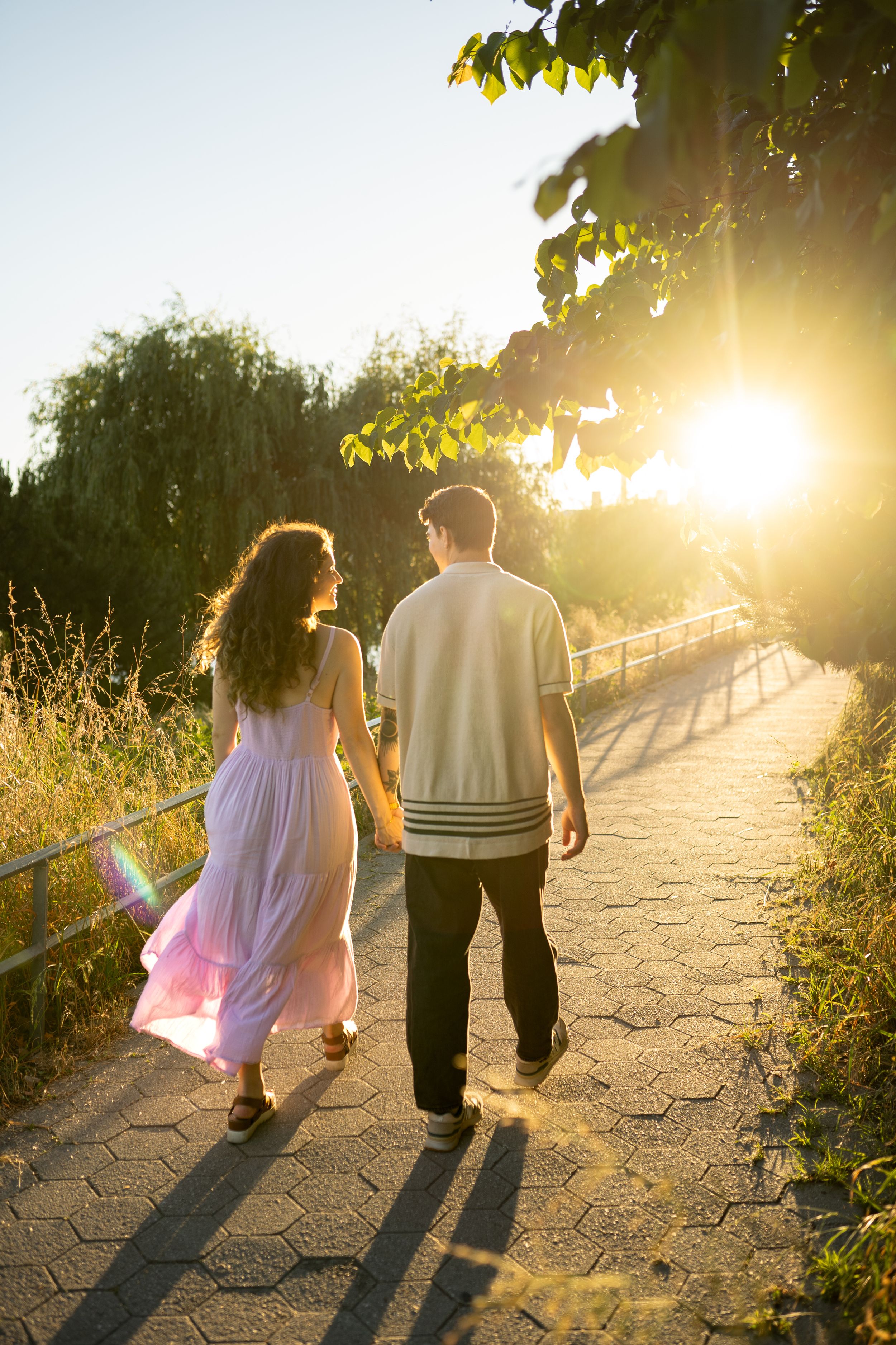 Two people walk hand in hand along a sunlit path with long shadows trailing behind them.