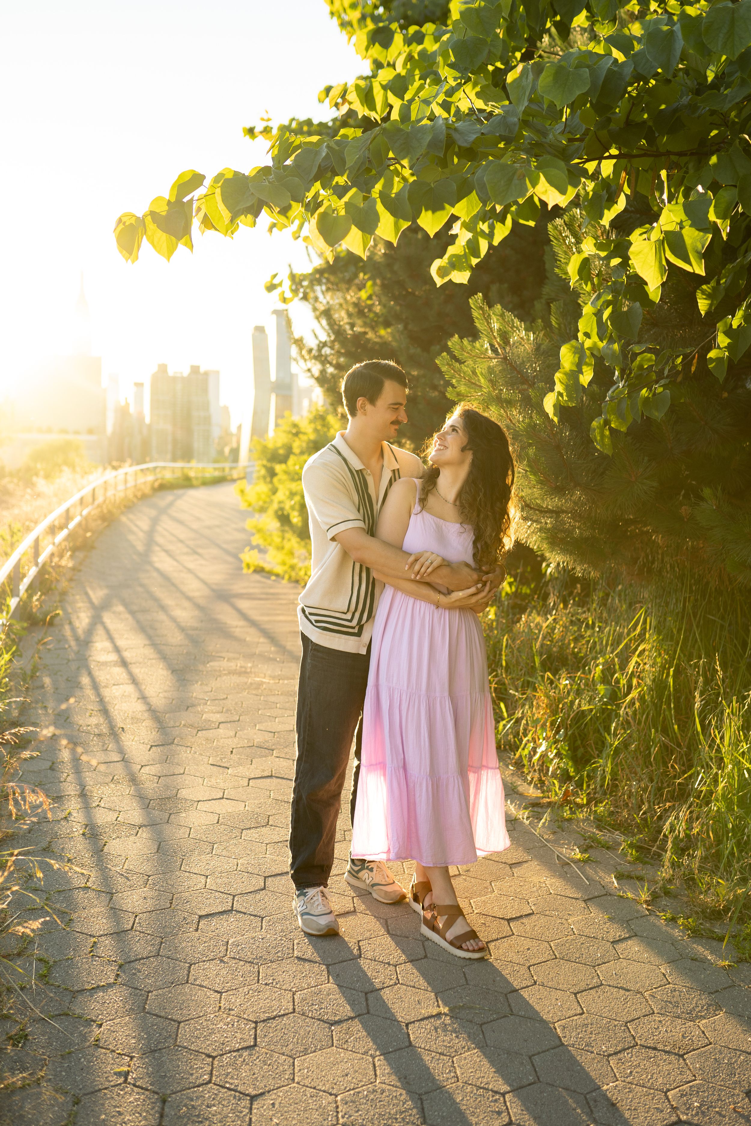 A romantic couple shares an intimate moment on a path during golden hour with the city skyline in the background.