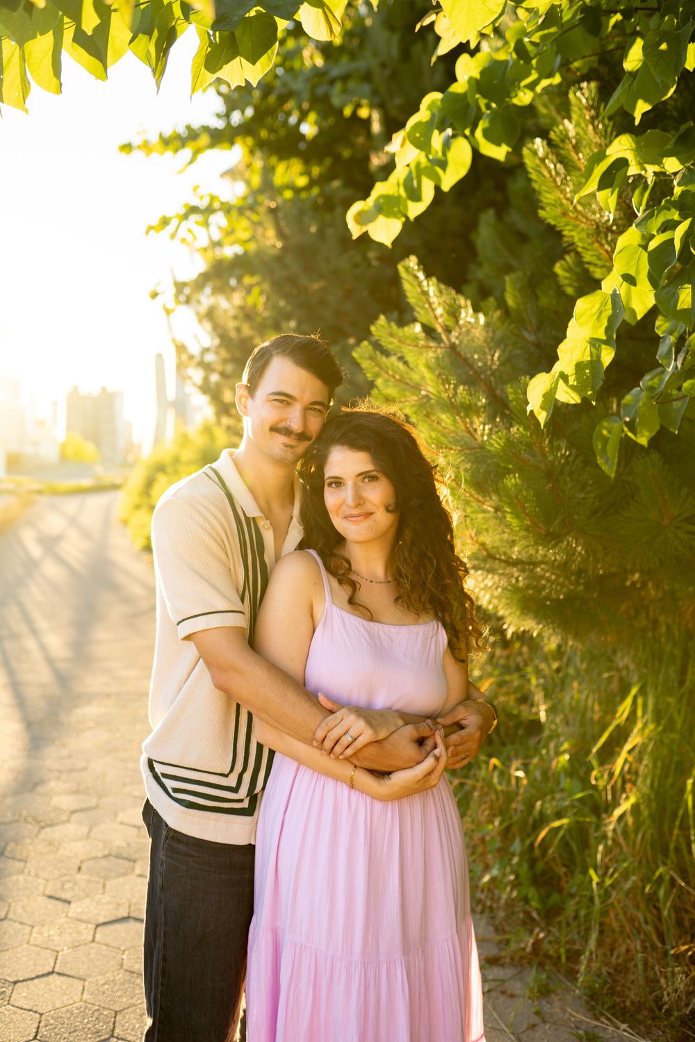 A loving couple embraces on a pathway during sunset with trees and city buildings in the distance.